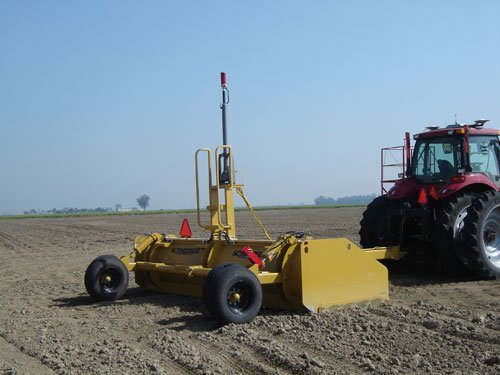 A yellow PFS open field leveler with its mast up, hitched to a red tractor on bare ground