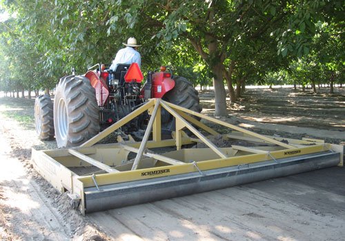 A grower on a red tractor pulling a yellow V-Blade orchard leveler down a shaded orchard row