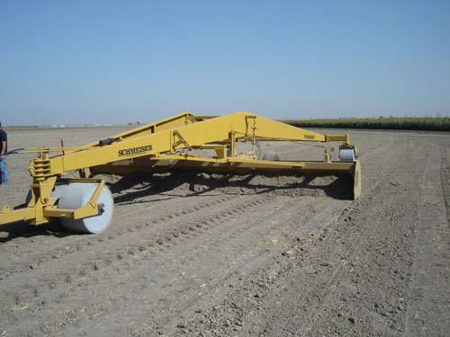 A yellow Smooth an’ Roll with its long leveling frame and white roller on freshly worked ground