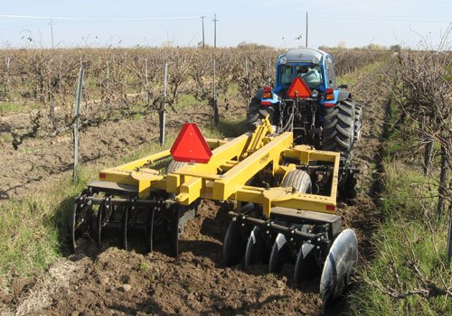 A yellow tandem disc turning soil between bare grapevines