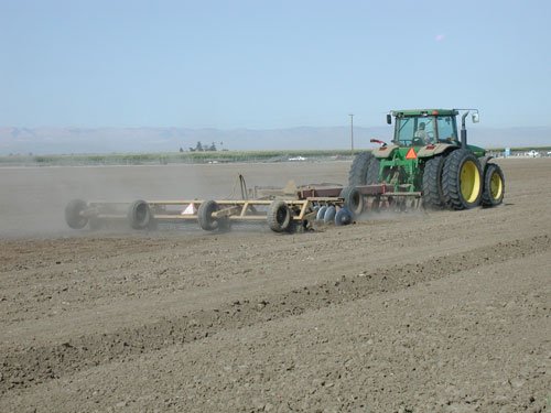 A green tractor pulling a long Till an’ Pak across a wide flat field under a hazy Valley sky