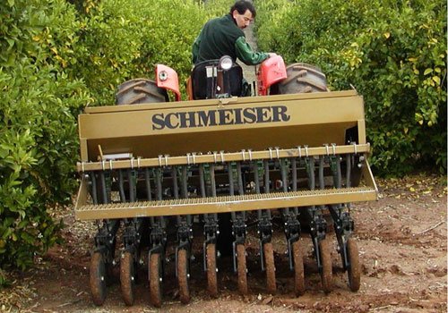 A Series 98 cover crop drill with SCHMEISER lettered on its hopper, pulled between citrus trees